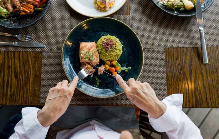 A person eating a dish of salmon, a piece of green rice, and mixed vegetables, surrounded by other dishes on the table.